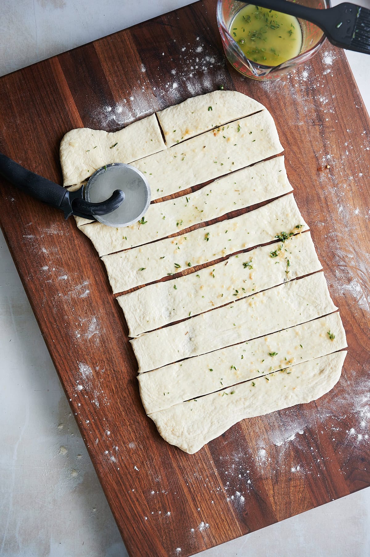 Rectangular sheet of dough on a floured wooden board, cut into strips with a pizza cutter, sprinkled with herbs, and a bowl of egg wash nearby.