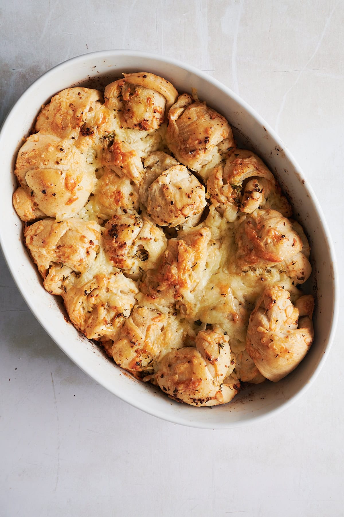 An oval baking dish filled with golden, baked pull-apart bread rolls topped with melted cheese and herbs.