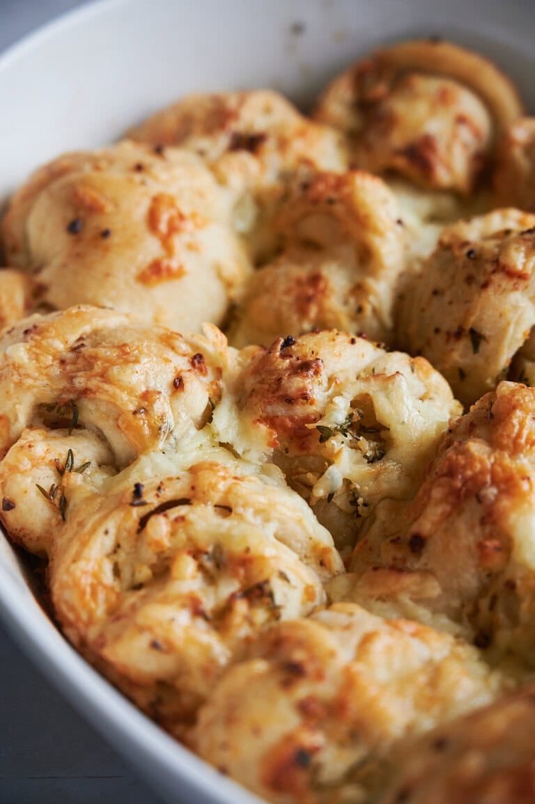 Golden brown cheesy garlic bread rolls in a white baking dish, with melted cheese and herbs visible on the flaky, baked surface.