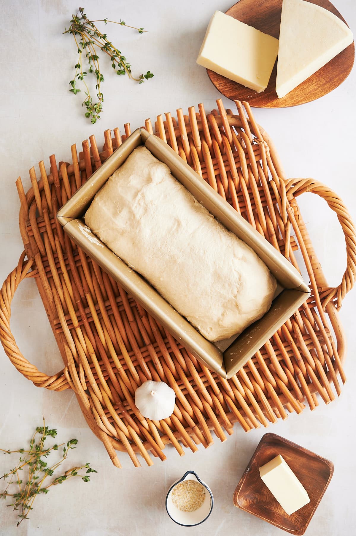 A loaf of unbaked bread dough in a rectangular pan sits on a wicker tray, surrounded by butter, garlic, cheese, and herbs.