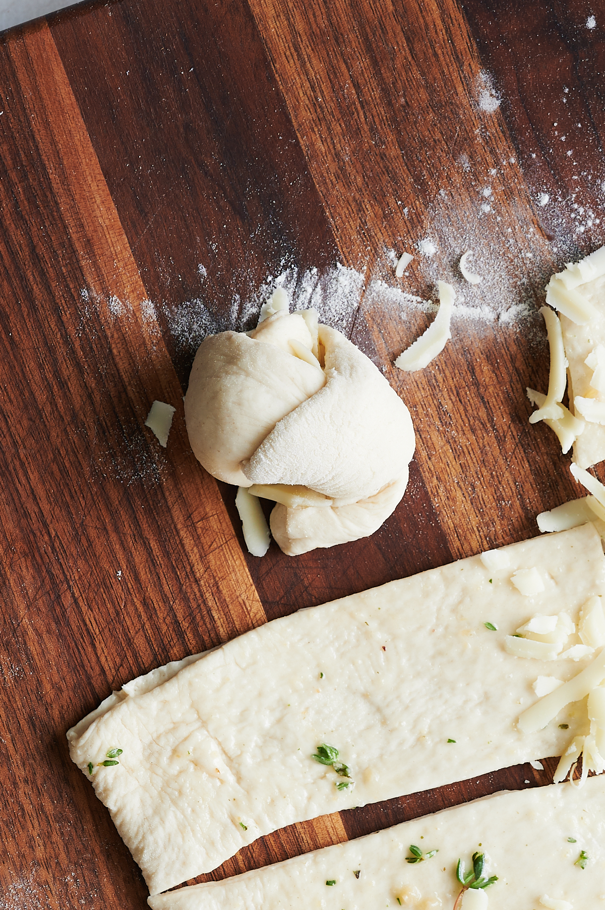 A ball of raw dough stuffed with cheese sits on a floured wooden surface next to a strip of dough topped with herbs and grated cheese.