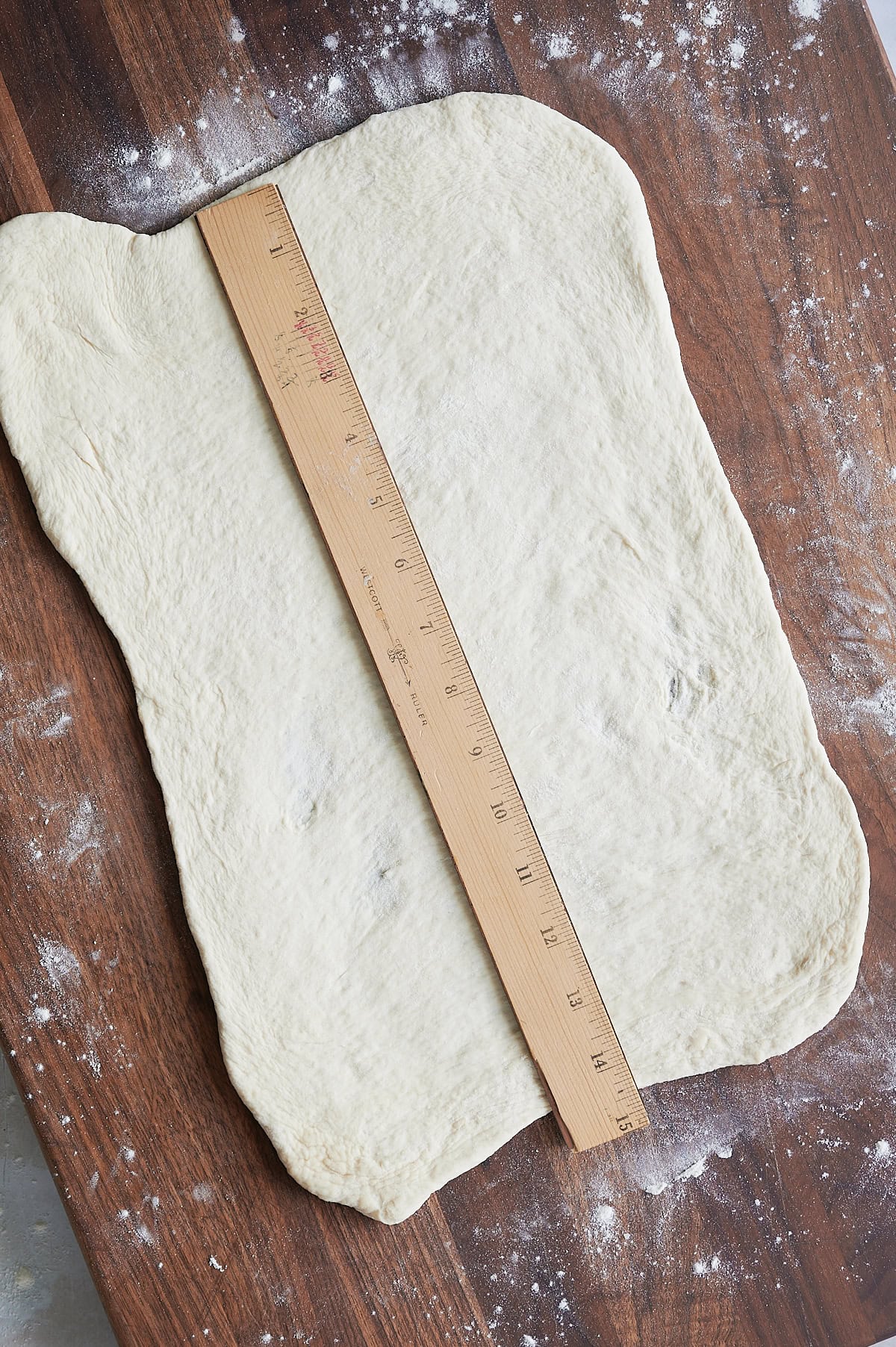 A wooden ruler measures the length of a rectangular sheet of dough on a floured wooden surface.