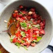 A gray green bowl of bruschetta chopped tomatoes, basil, and oil with a spoon, next to a small bowl of salt on a white surface.