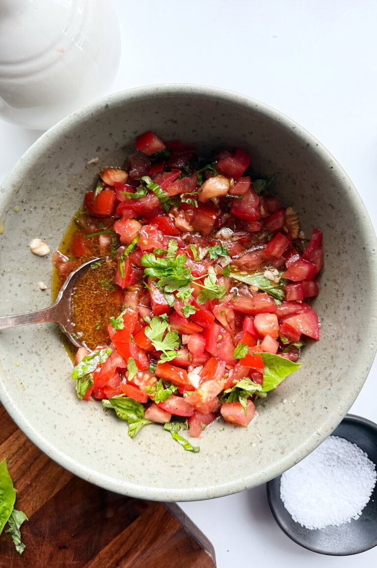 A gray green bowl of bruschetta chopped tomatoes, basil, and oil with a spoon, next to a small bowl of salt on a white surface.