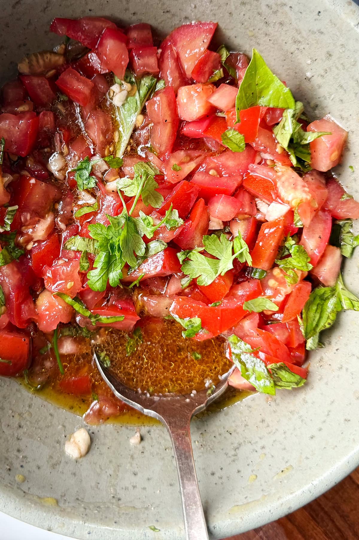 A bowl of bruschetta with chopped tomatoes, fresh herbs, and minced garlic mixed with olive oil, with a metal spoon resting inside.