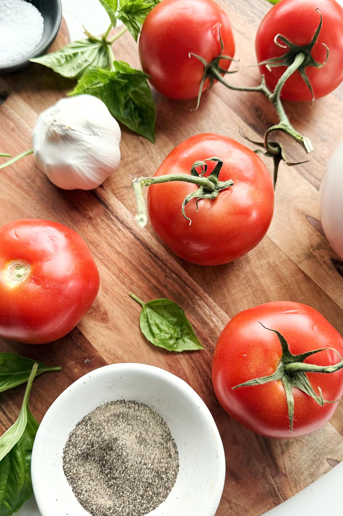 Fresh tomatoes, basil leaves, garlic, a bowl of ground black pepper, and a dish of salt arranged on a wooden surface.