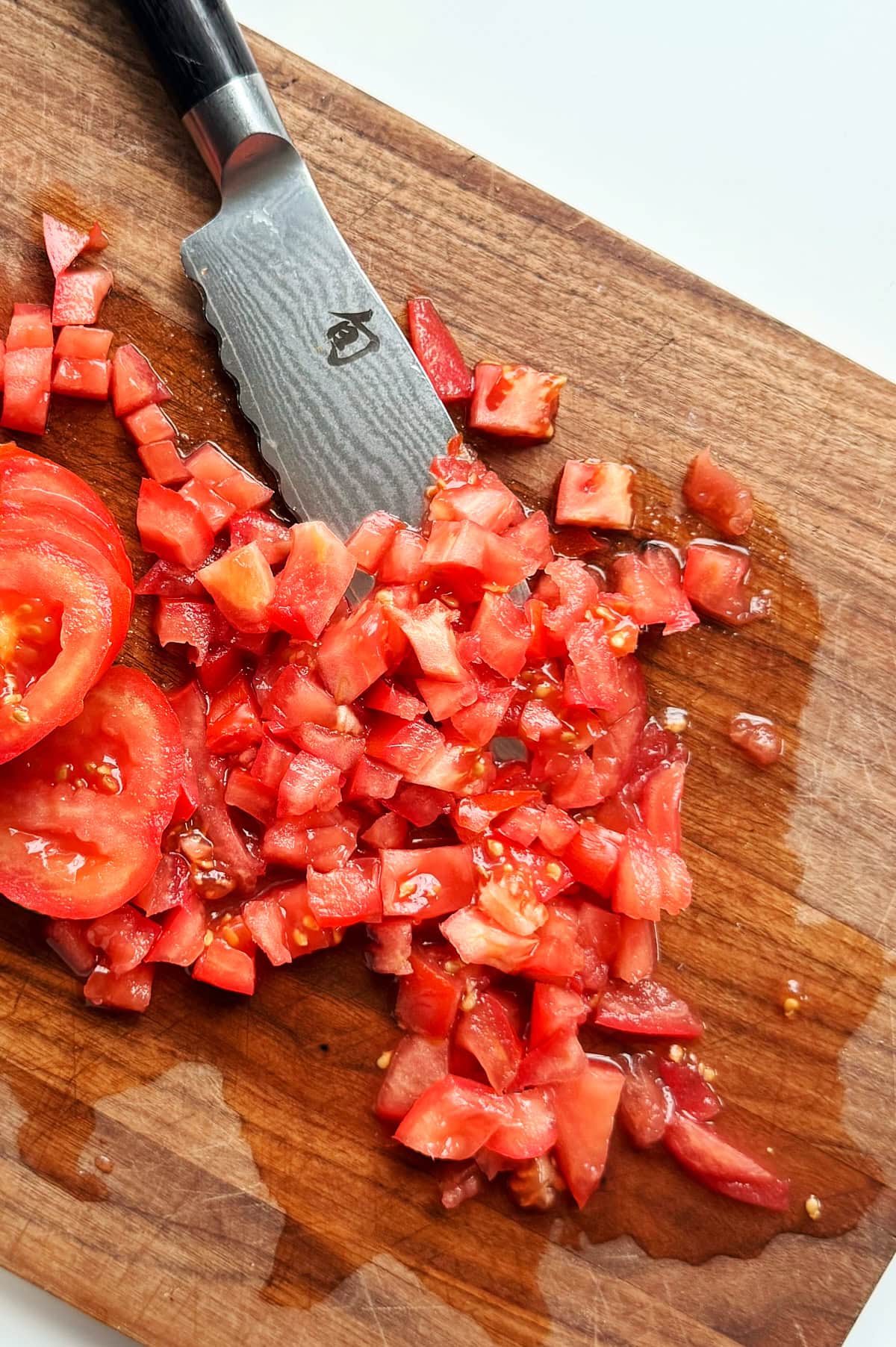 A kitchen knife and diced tomatoes are on a wooden cutting board, with tomato juices visible.