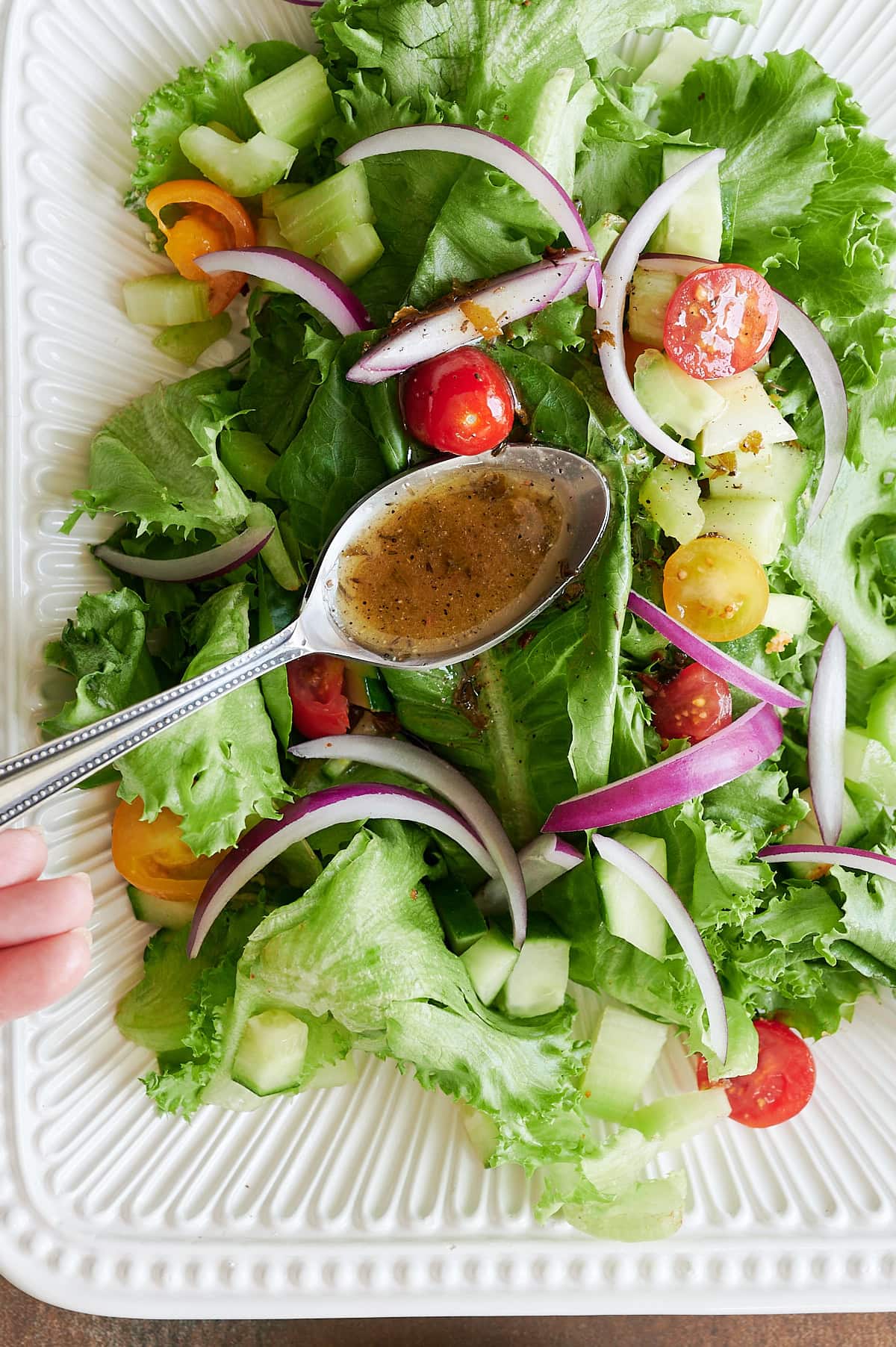 A hand holding a spoon drizzles Italian salad dressing over a green salad with lettuce, cherry tomatoes, red onion, and cucumber on a white plate.