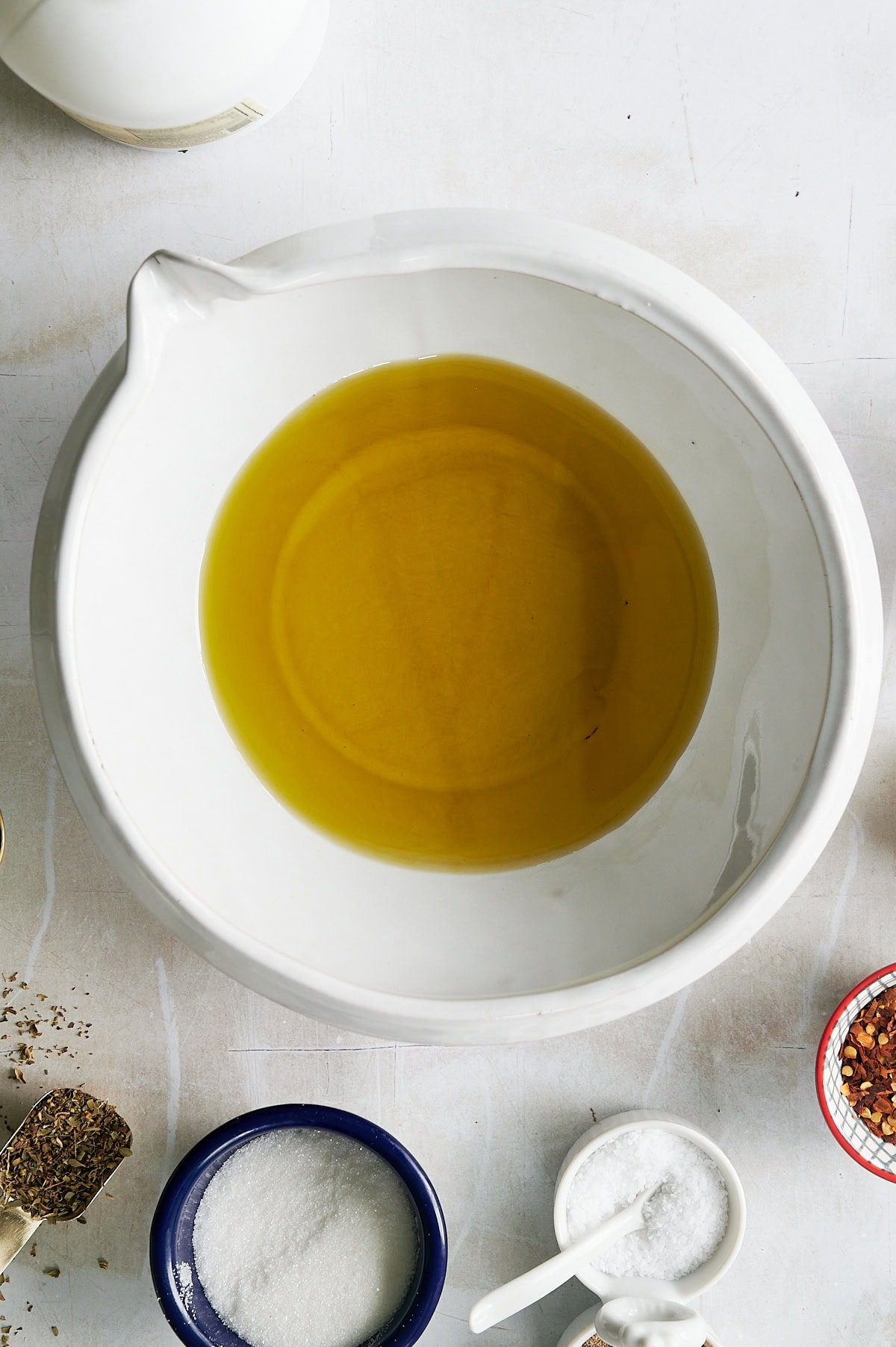 A white bowl filled with olive oil sits on a white surface, surrounded by bowls and spoons containing salt, sugar, spices, and seasonings.
