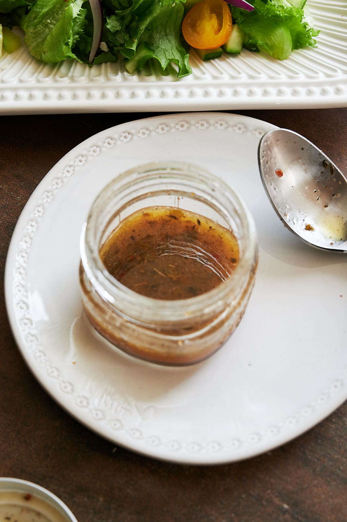 A glass jar with Italian salad dressing sits on a white plate next to a metal spoon, with a plate of salad partially visible in the background.