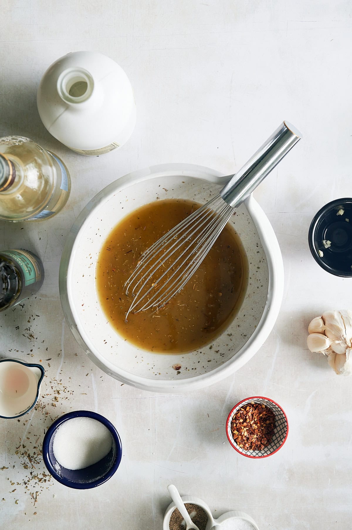A mixing bowl with a whisk mixing salad dressing surrounded by ingredients including a jug, spices, sugar, and ginger on a light countertop.