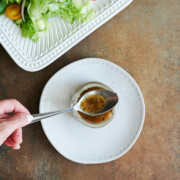 A hand holds a spoon over a small jar of salad dressing on a white plate, with a green salad and a bowl of grated cheese nearby.