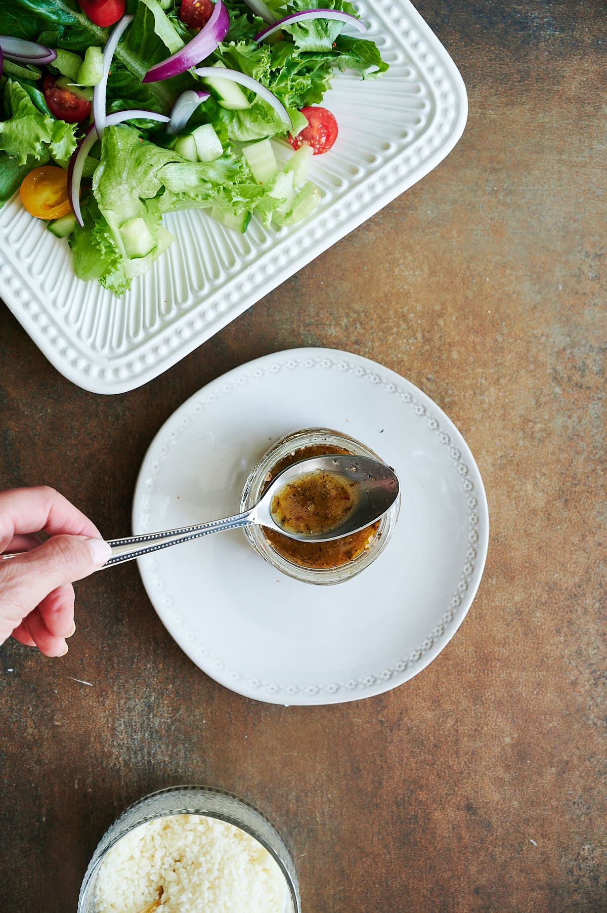 A hand holds a spoon over a small jar of salad dressing on a white plate, with a green salad and a bowl of grated cheese nearby.