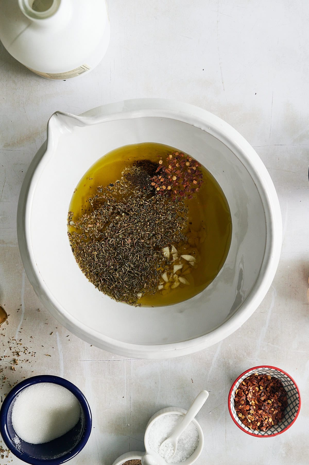 A white bowl containing olive oil, dried herbs, chopped garlic, and red pepper flakes for Italian salad dressing sits on a white surface with salt, sugar, and a bottle also visible nearby.