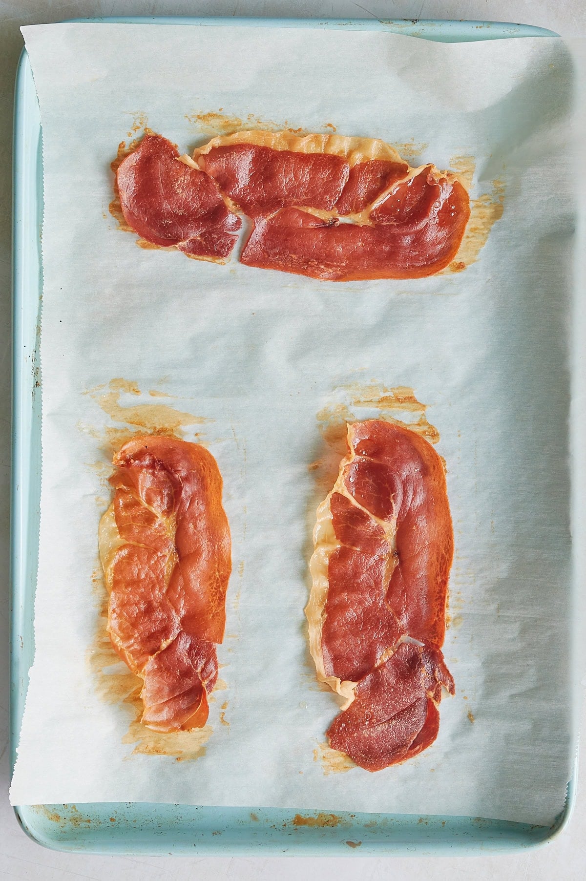Three slices of prosciutto are baked on a parchment-lined baking sheet, with two at the bottom and one at the top.