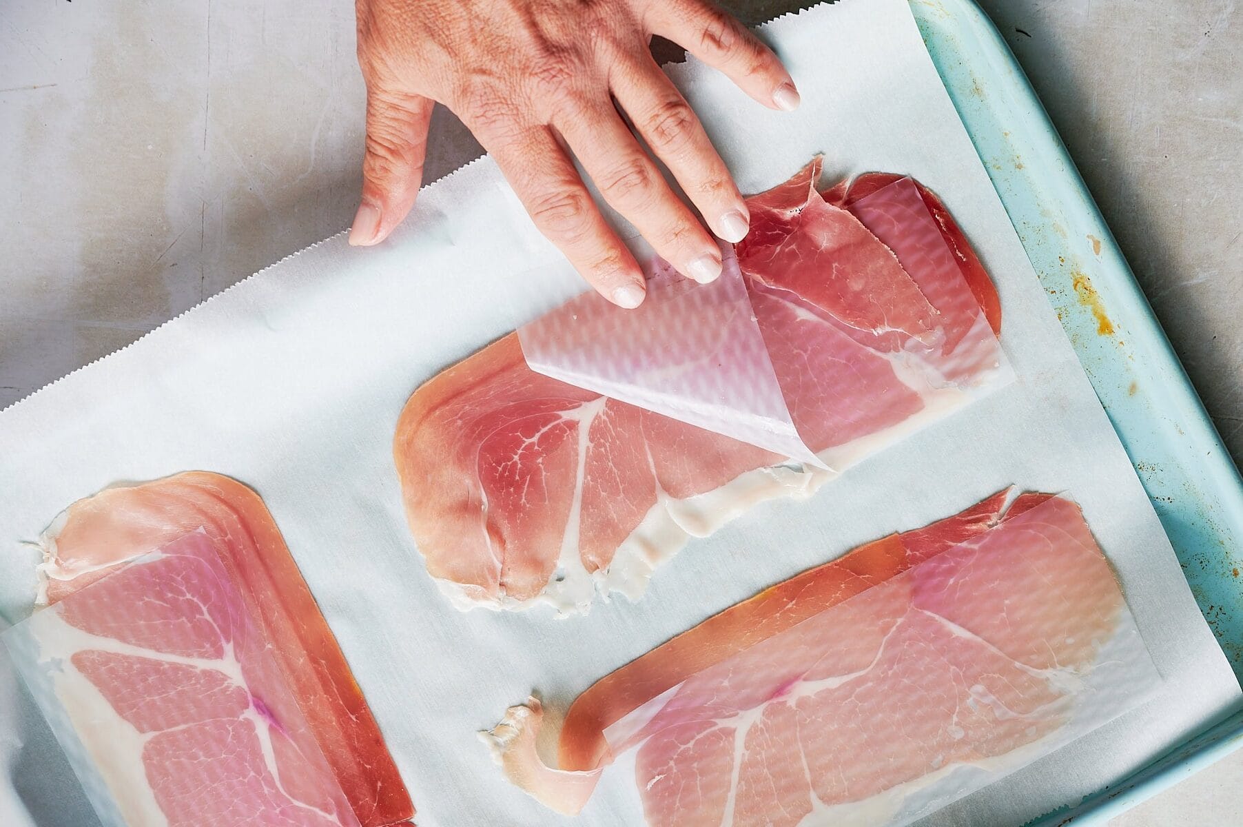Hand peeling back parchment paper from thin slices of prosciutto arranged on a baking sheet lined with parchment paper.