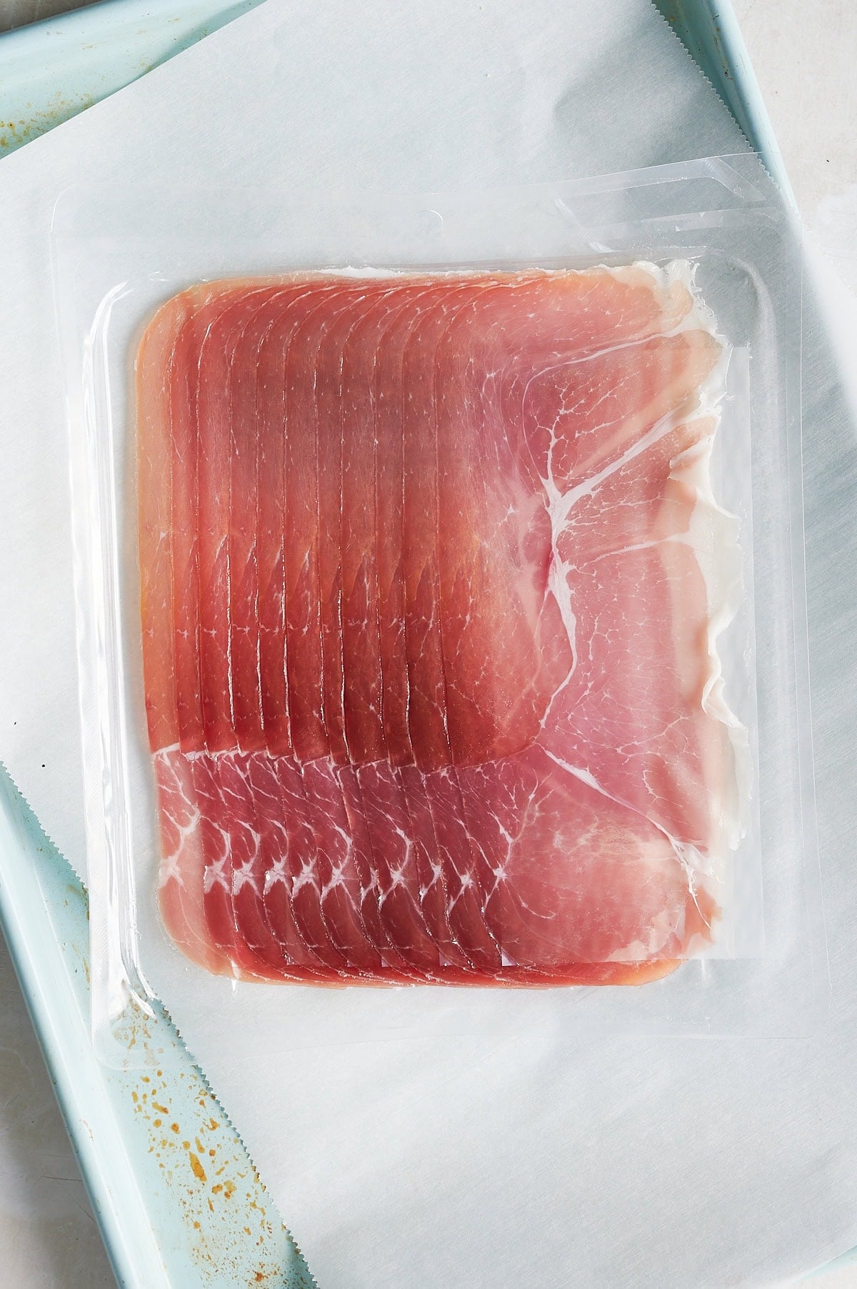 A vacuum-sealed package of thinly sliced prosciutto on a white tray lined with parchment paper.
