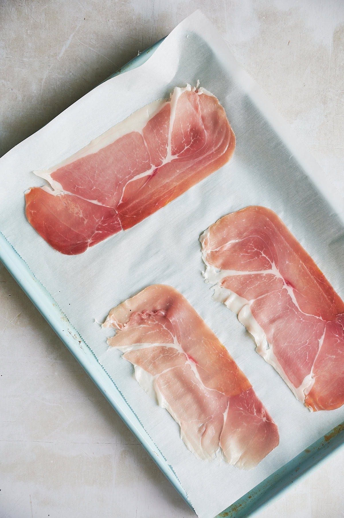 Three slices of prosciutto are arranged on a parchment-lined baking sheet.