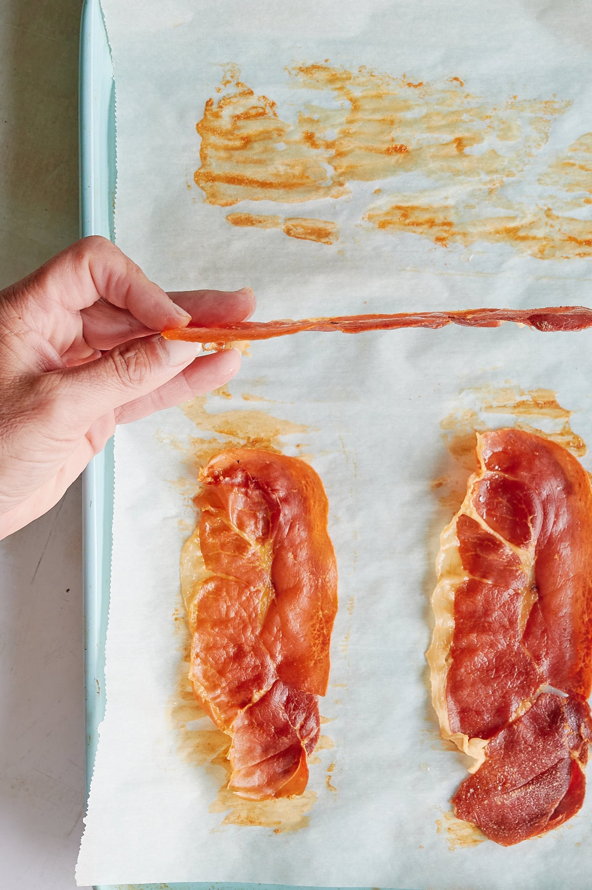 A hand holds a thin, crispy strip of prosciutto above a baking tray lined with parchment paper and two baked prosciutto slices.