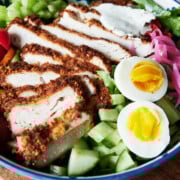 A bowl of sliced breaded chicken cutlet, hard-boiled egg halves, cucumber, lettuce, celery, tomatoes, pickled onions, and a dollop of dressing.