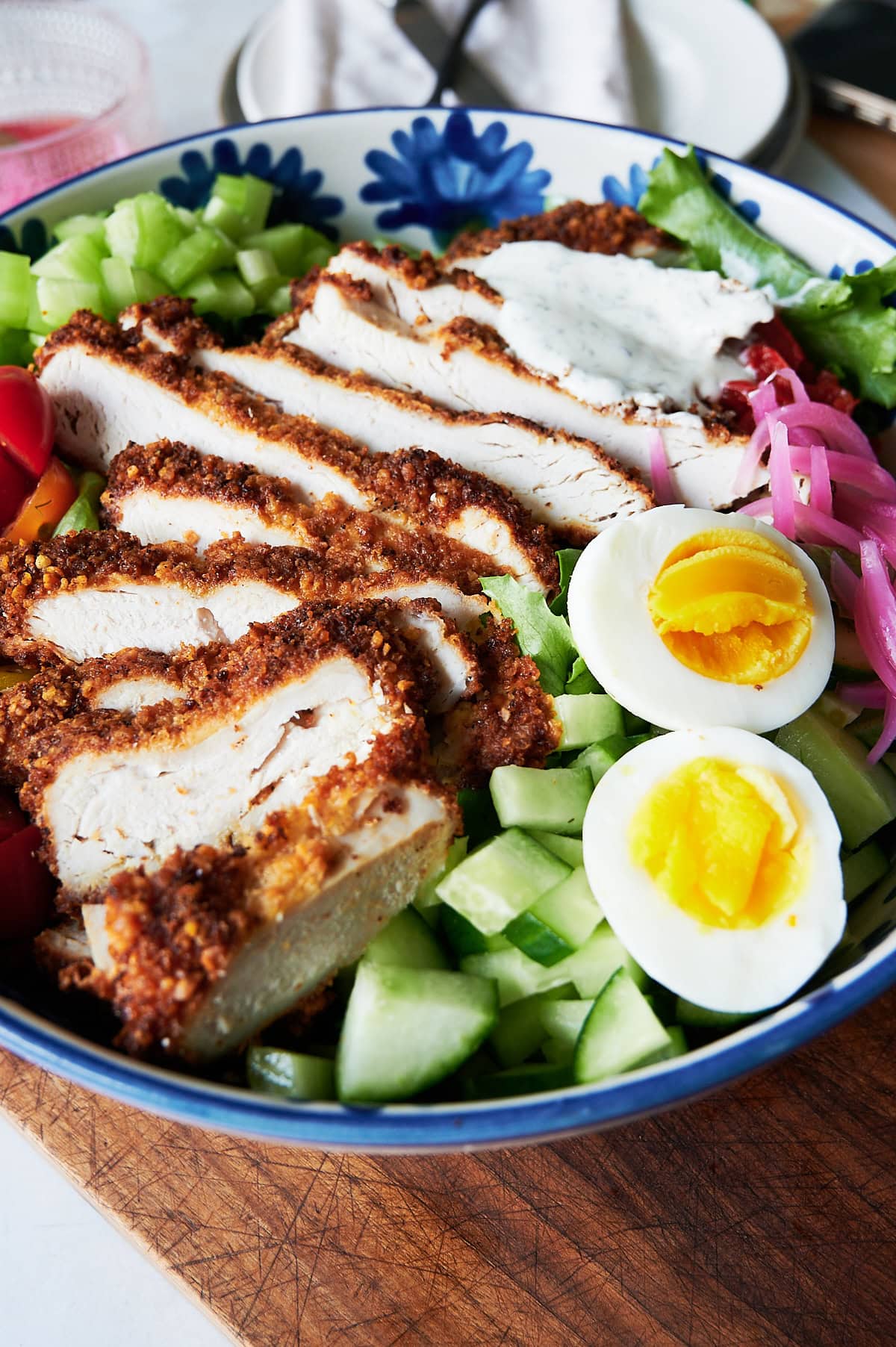 A bowl of sliced breaded chicken cutlet, hard-boiled egg halves, cucumber, lettuce, celery, tomatoes, pickled onions, and a dollop of dressing.