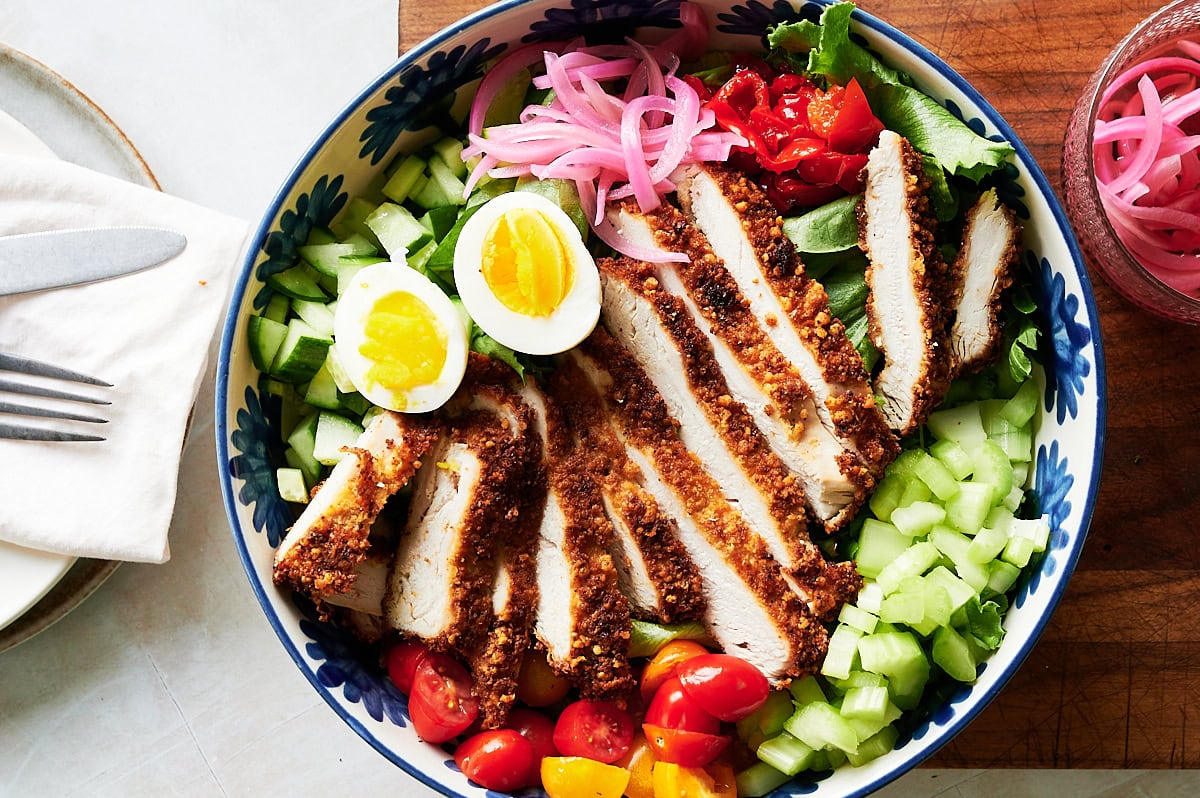 Looking down at a salad of sliced breaded fried chicken breast, halved boiled egg, cherry tomatoes, celery, cucumbers, mixed greens, and pickled onions arranged neatly.