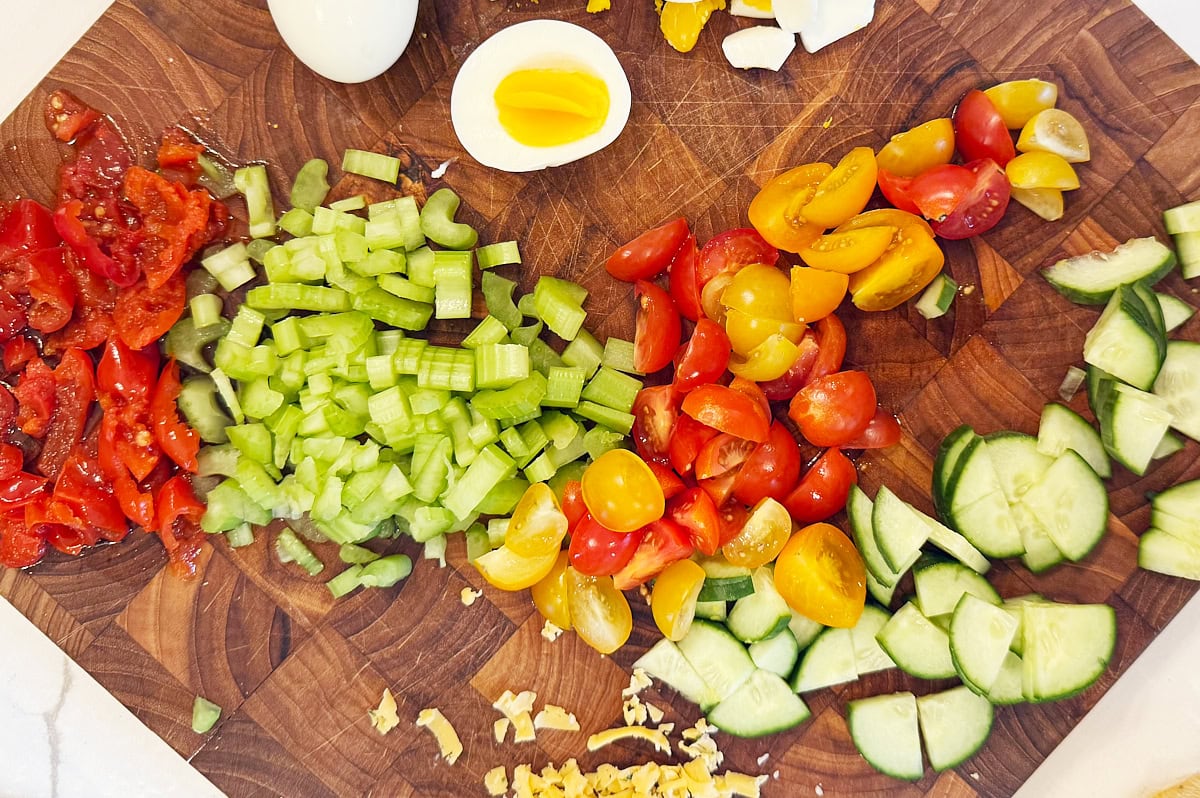 Chopped tomatoes, celery, cherry tomatoes, cucumbers, and hard-boiled egg pieces arranged on a wooden cutting board.