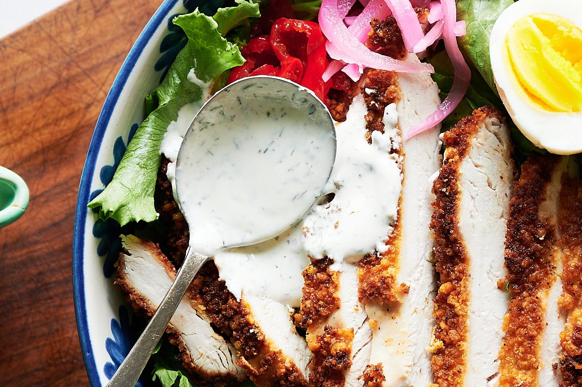 A bowl with sliced breaded chicken, leafy greens, pickled onions, red peppers, half a boiled egg, and a spoonful of creamy dressing.