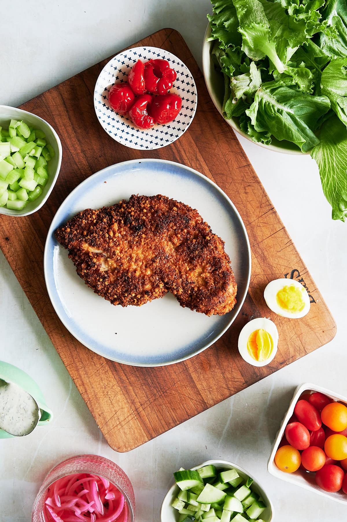 A plate with a breaded, fried chicken breast on a wooden board surrounded by bowls of lettuce, celery, cherry tomatoes, cucumber, red onions, salad dressing, an egg, and red peppers.