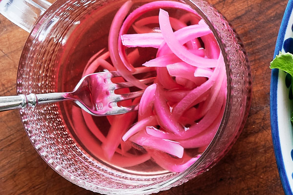 A glass bowl filled with sliced pickled red onions and a fork resting inside, placed on a wooden surface.