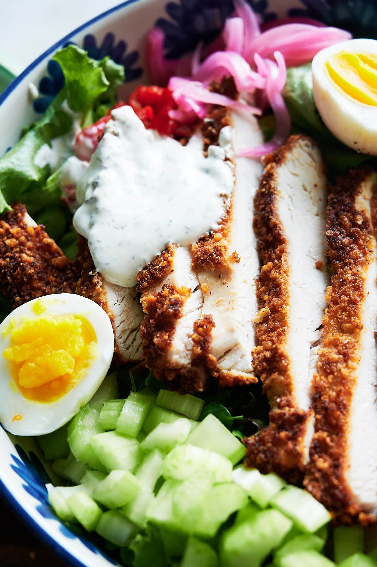 A salad bowl with sliced chicken Milanese, hard-boiled egg halves, chopped cucumber, greens, pickled onions, and a dollop of creamy dressing.