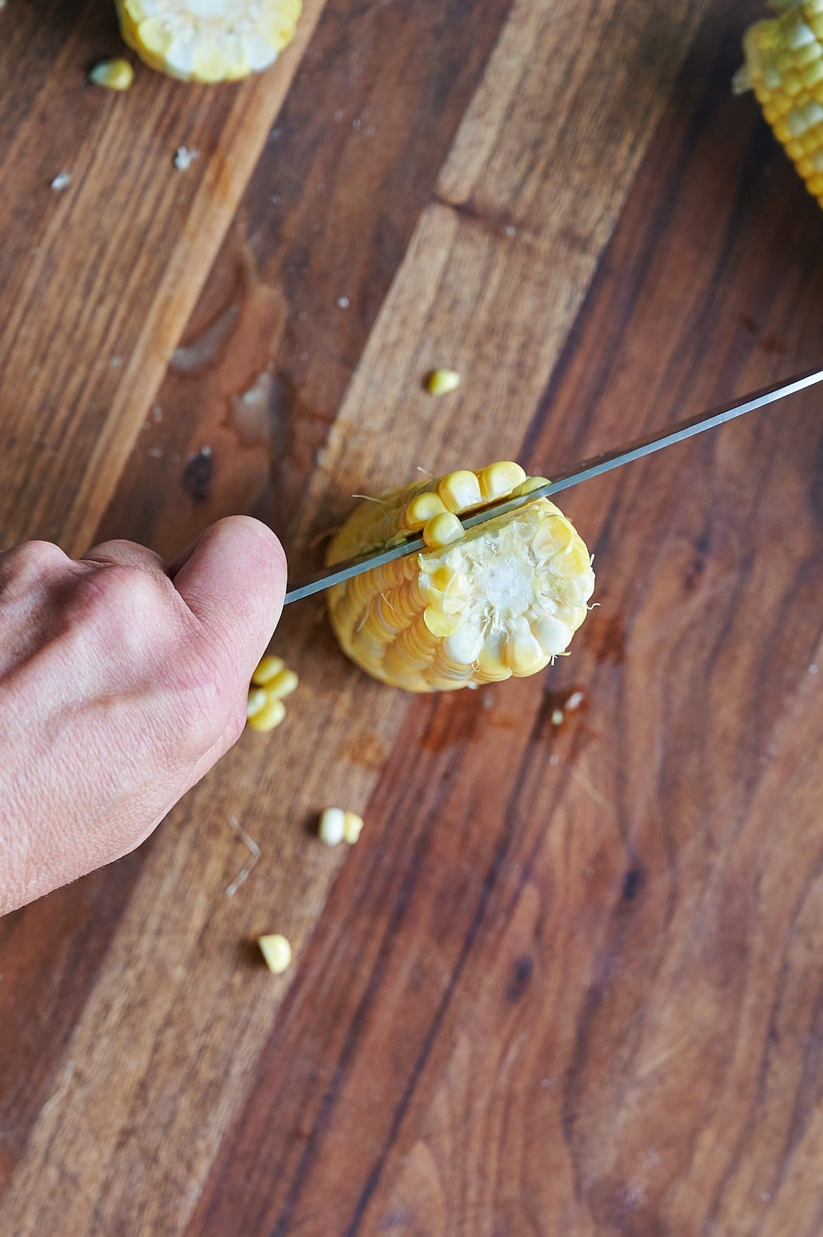 A hand holds an ear of corn upright on a wooden cutting board while a knife slices through the corn kernels off the cob.