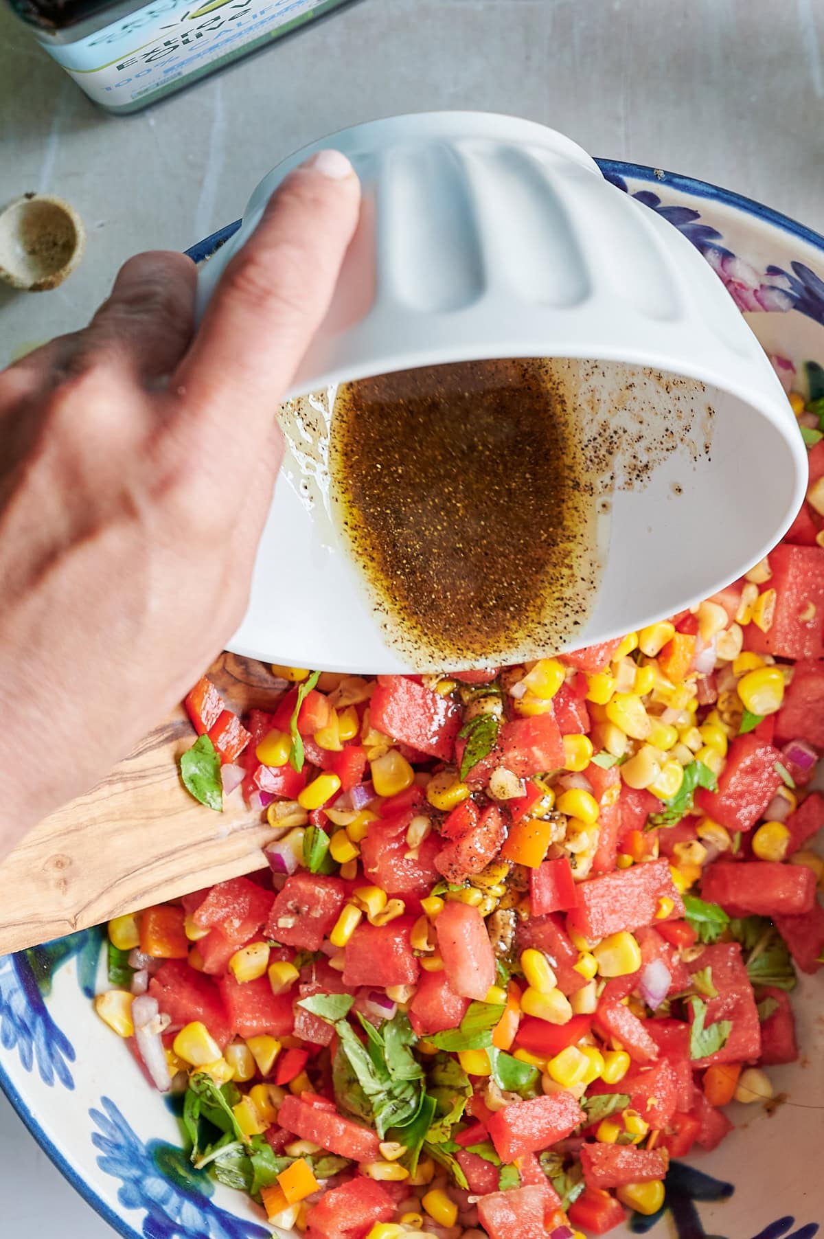 A hand pours a balsamic salad dressing from a white bowl onto a salad made of watermelon, corn, bell pepper, onion, and basil in a large dish.