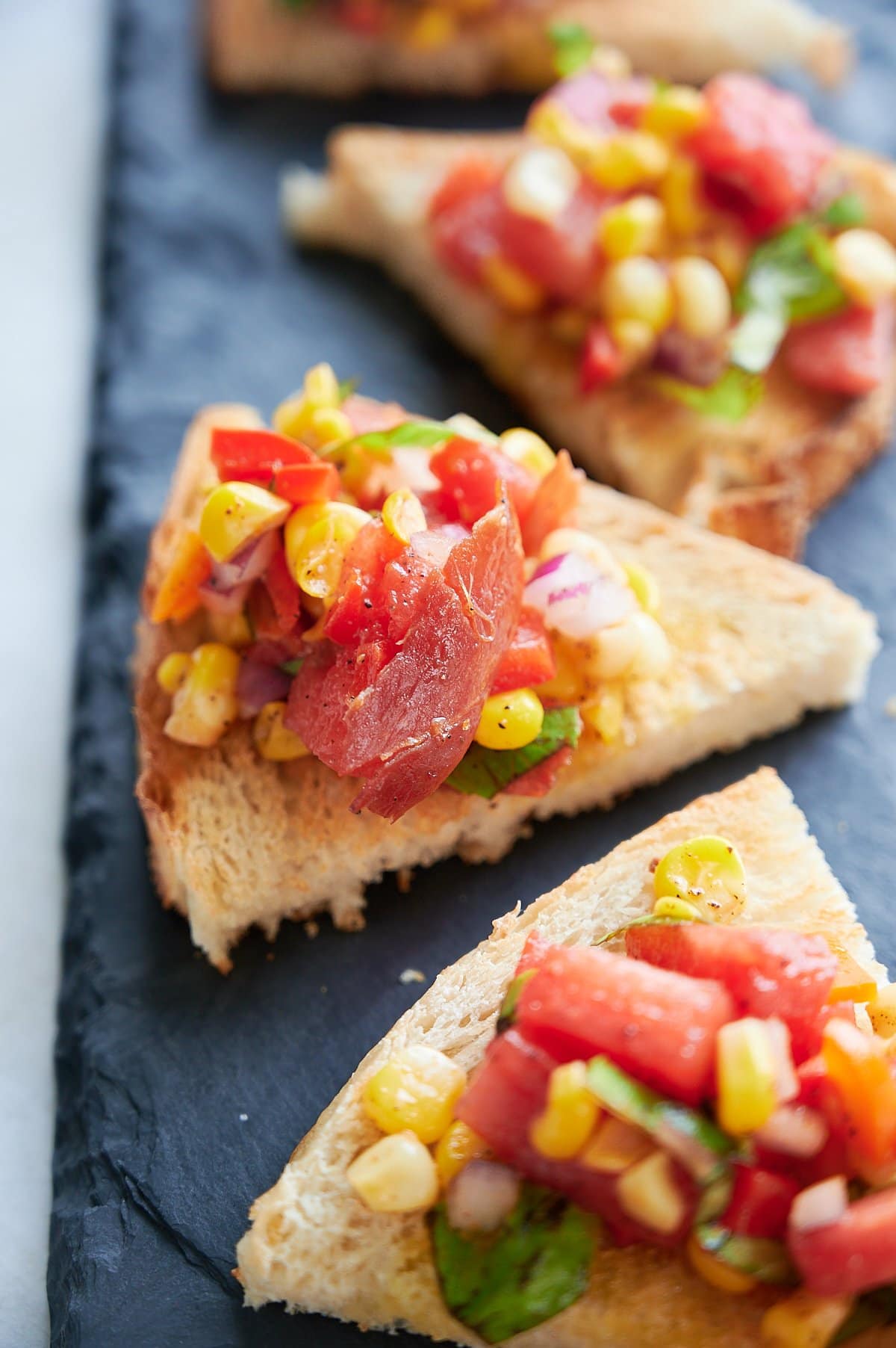 Triangular slices of toasted bread topped with a mixture corn bruschetta arranged on a dark slate serving tray.