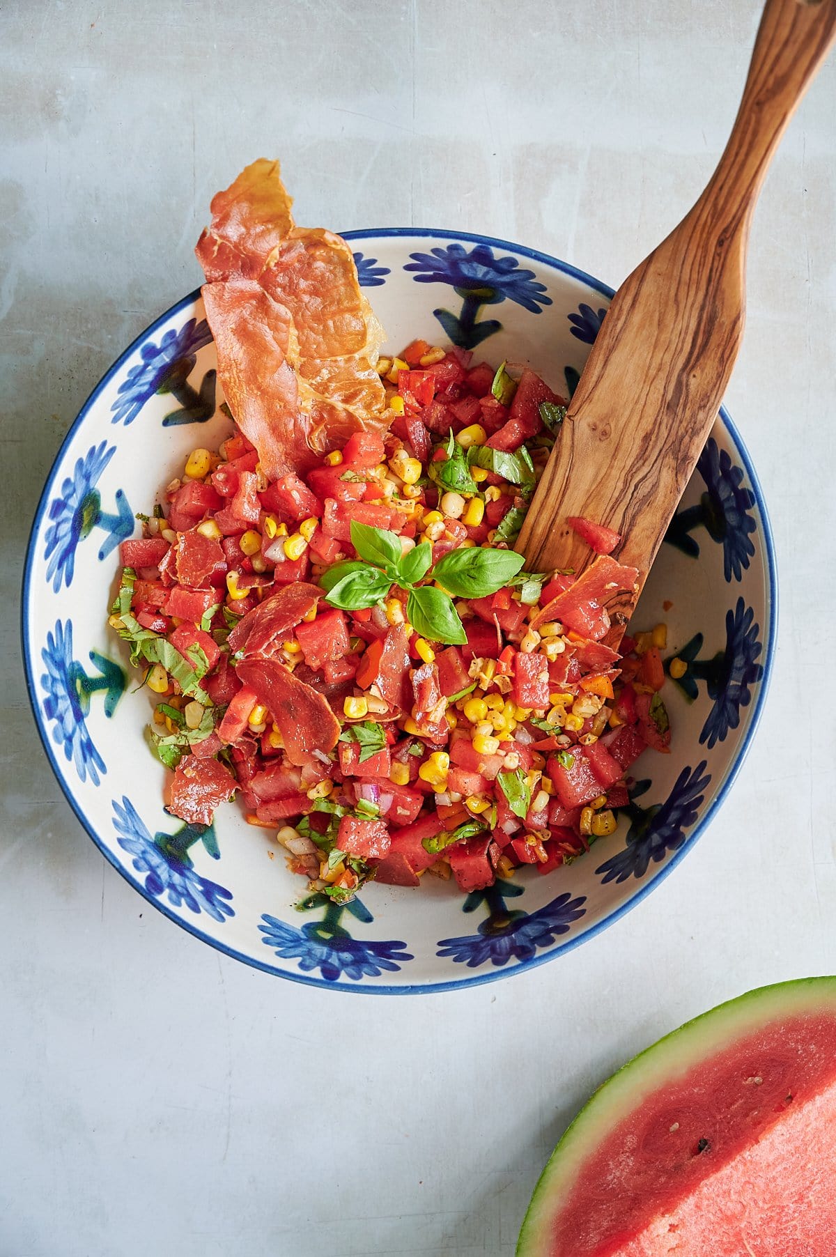 A bowl of watermelon salad with corn, fresh basil, and crispy prosciutto, served with a wooden spoon. A slice of watermelon is visible in the corner.