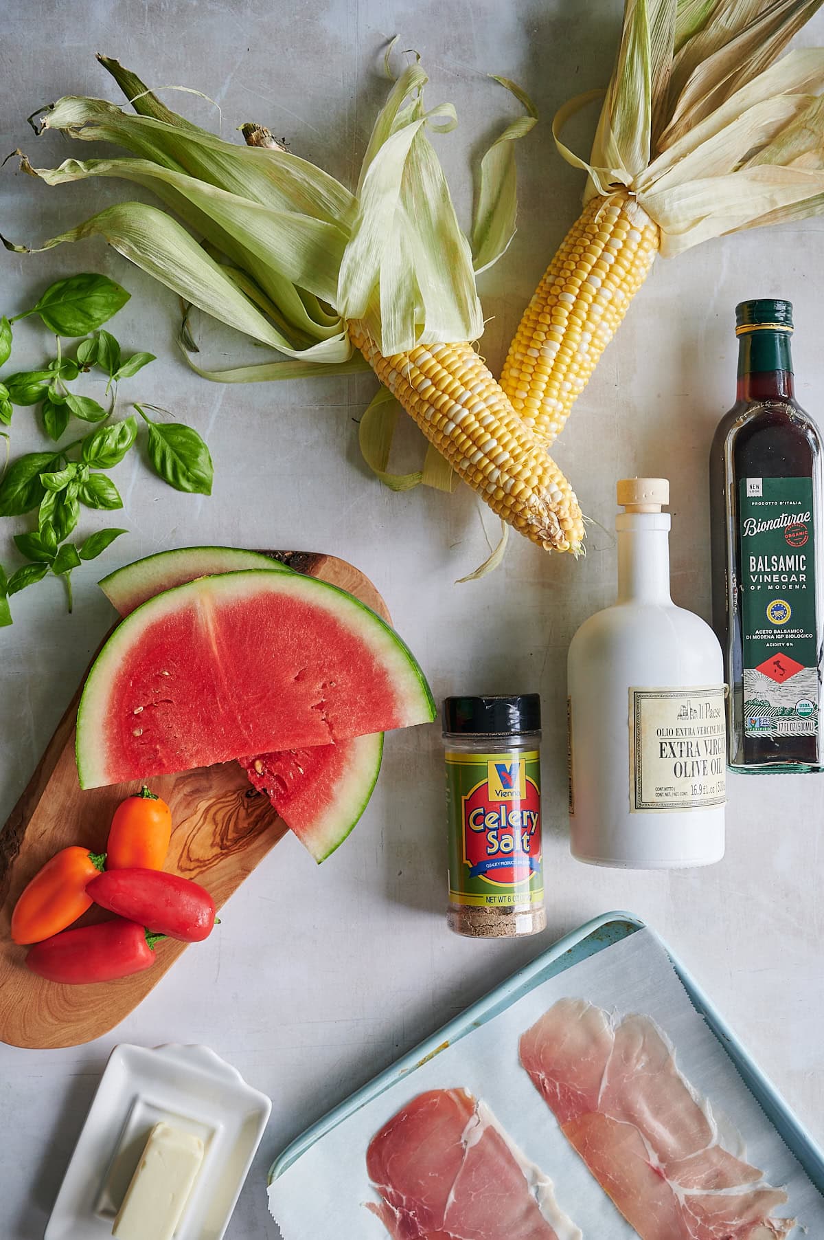 An arrangement of fresh corn, watermelon slices, basil, sweet peppers, prosciutto, butter, olive oil, balsamic vinegar, and celery salt on a light surface.