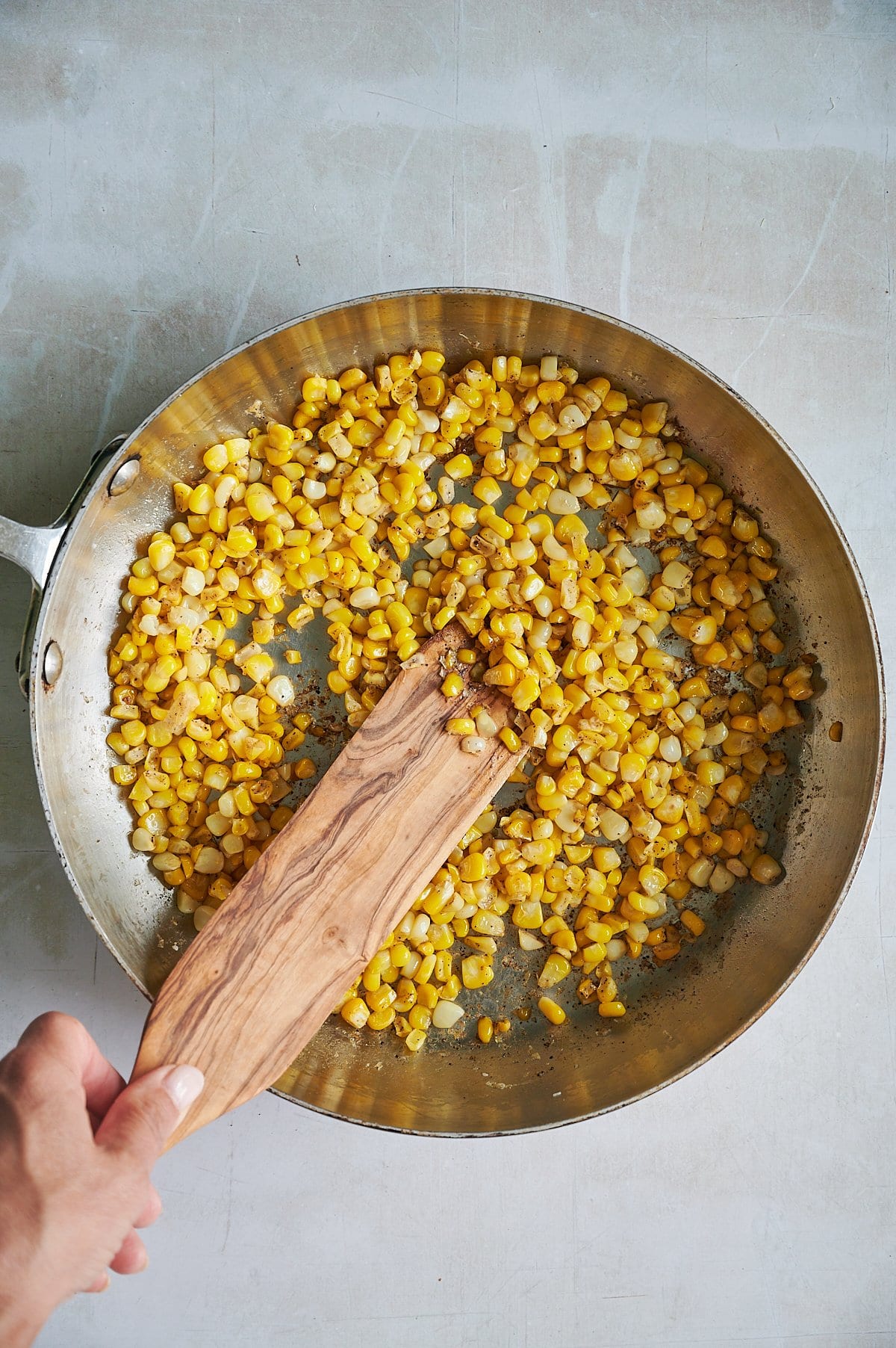 A hand stirs sautéed corn kernels in a metal pan with a wooden spatula on a light-colored surface.