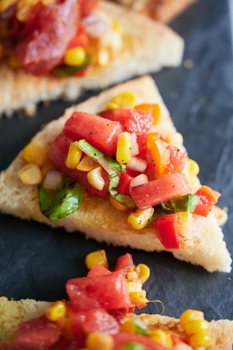 Close-up of toasted bread topped with diced tomatoes, corn, onion, and basil on a dark surface.