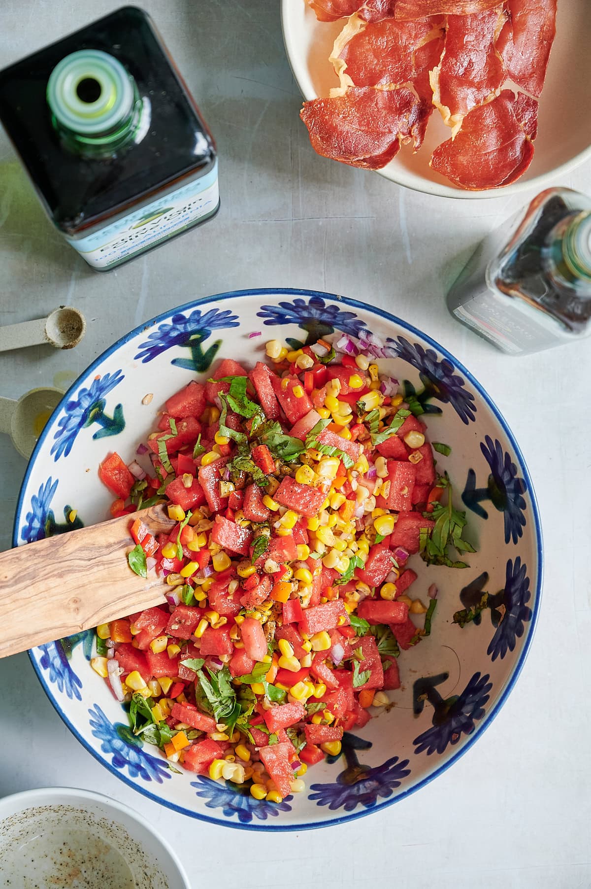 A bowl of watermelon, corn, and herb salad sits on a table next to bottles of olive oil and vinegar, with a separate bowl of prosciutto.