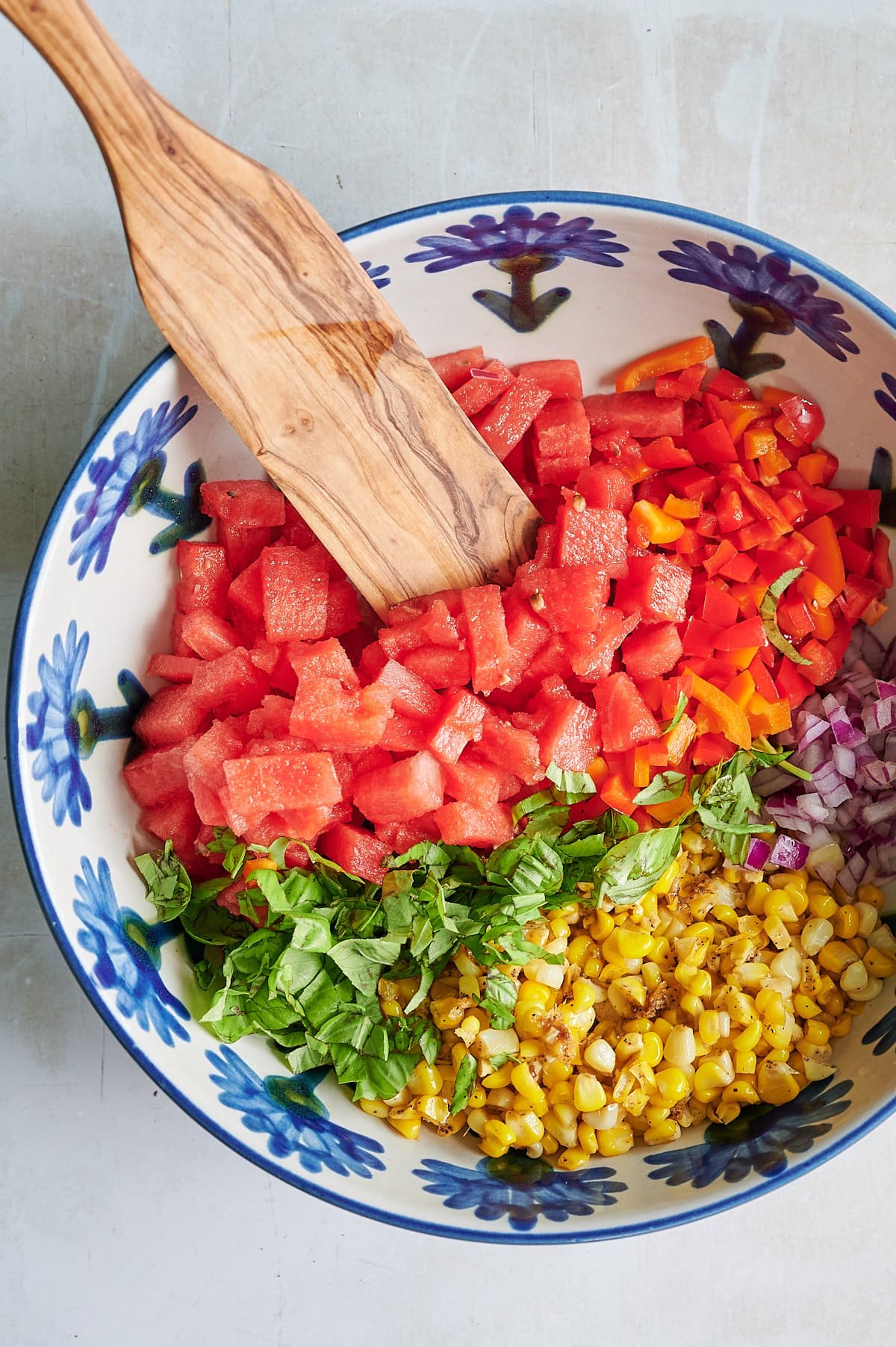 A bowl contains diced watermelon, chopped red and yellow bell peppers, red onion, corn, and chopped basil, with a wooden spatula resting on the edge.