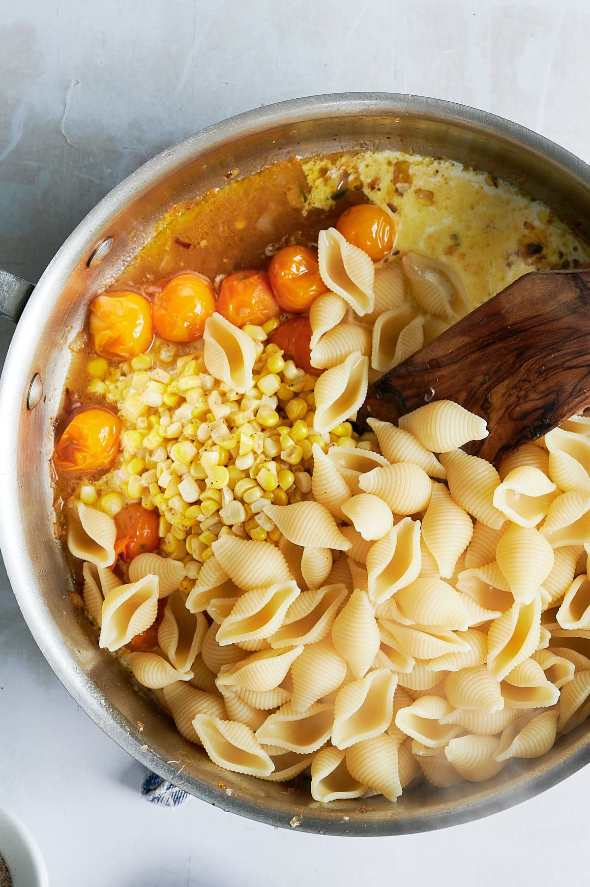 A wooden paddle stirring cooked pasta shells into sauteed cherry tomatoes, corn kernels and a creamy sauce.