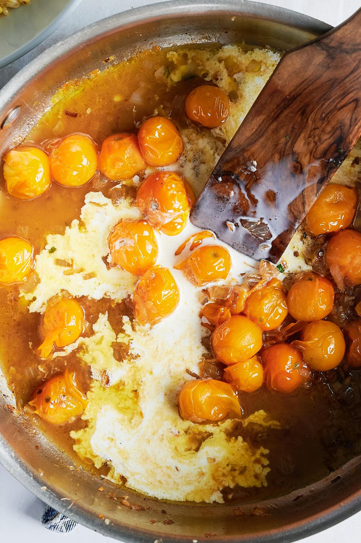 A metal pan with yellow-orange cherry tomatoes cooking in a yellow sauce, partially mixed with cream, and a wooden spatula resting along the side.