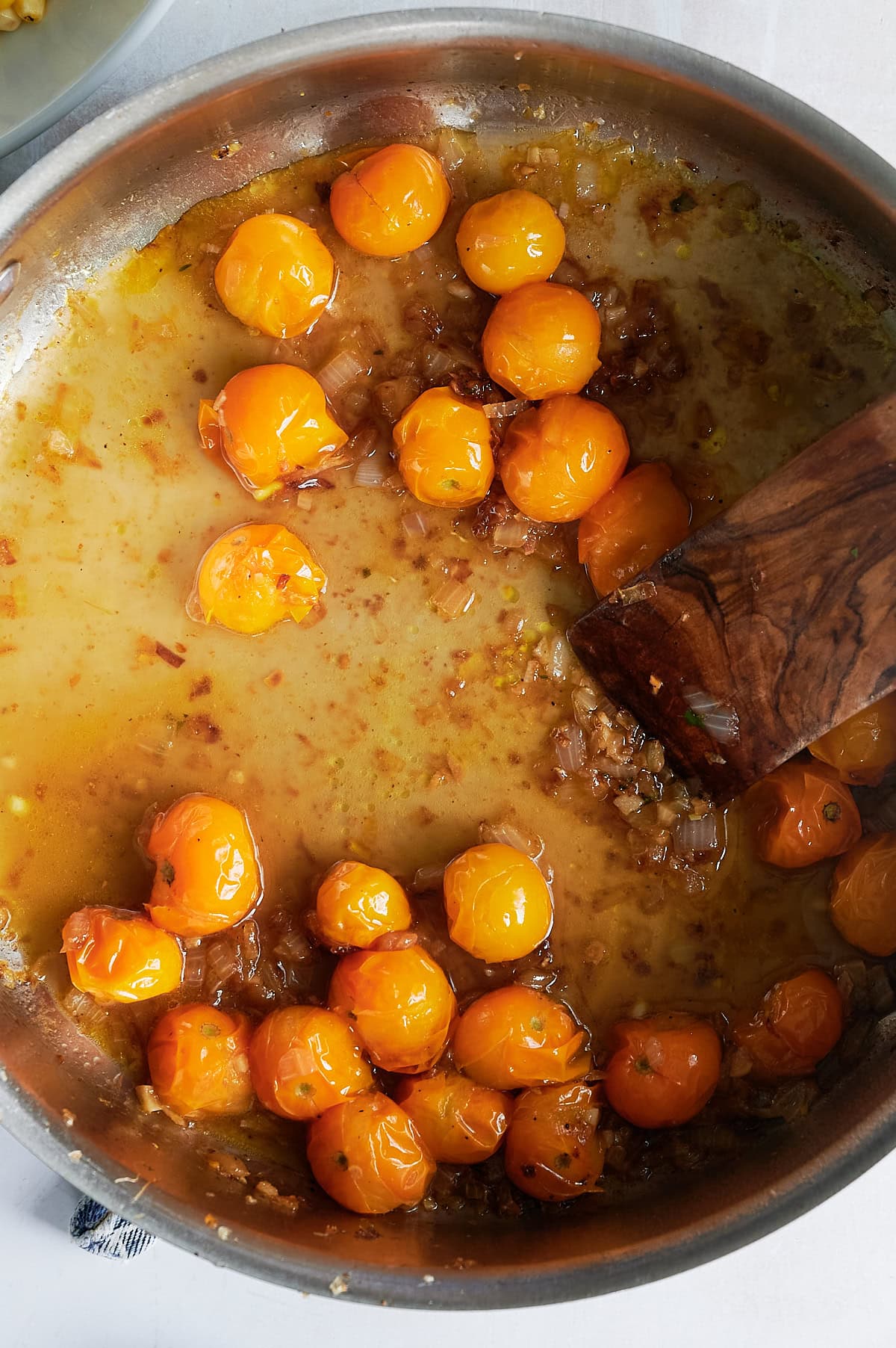A stainless steel pan of yellow cherry tomatoes being deglazed with a wooden spatula.