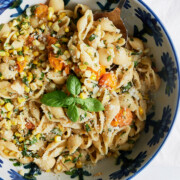 A blue floral bowl of shell pasta with corn, cherry tomatoes, herbs, grated cheese, and a fresh basil garnish, with a serving spoon placed in the bowl.