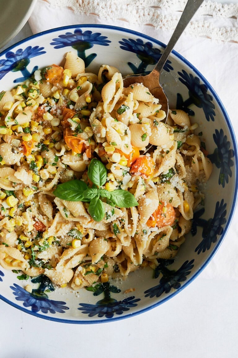 A blue floral bowl of shell pasta with corn, cherry tomatoes, herbs, grated cheese, and a fresh basil garnish, with a serving spoon placed in the bowl.