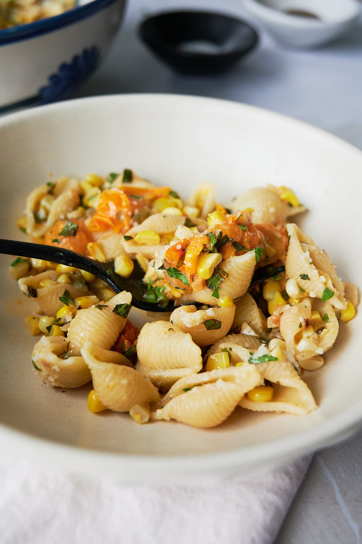 A bowl of shell pasta mixed with corn, chopped tomatoes, herbs, and a black spoon resting in a white pasta bowl.