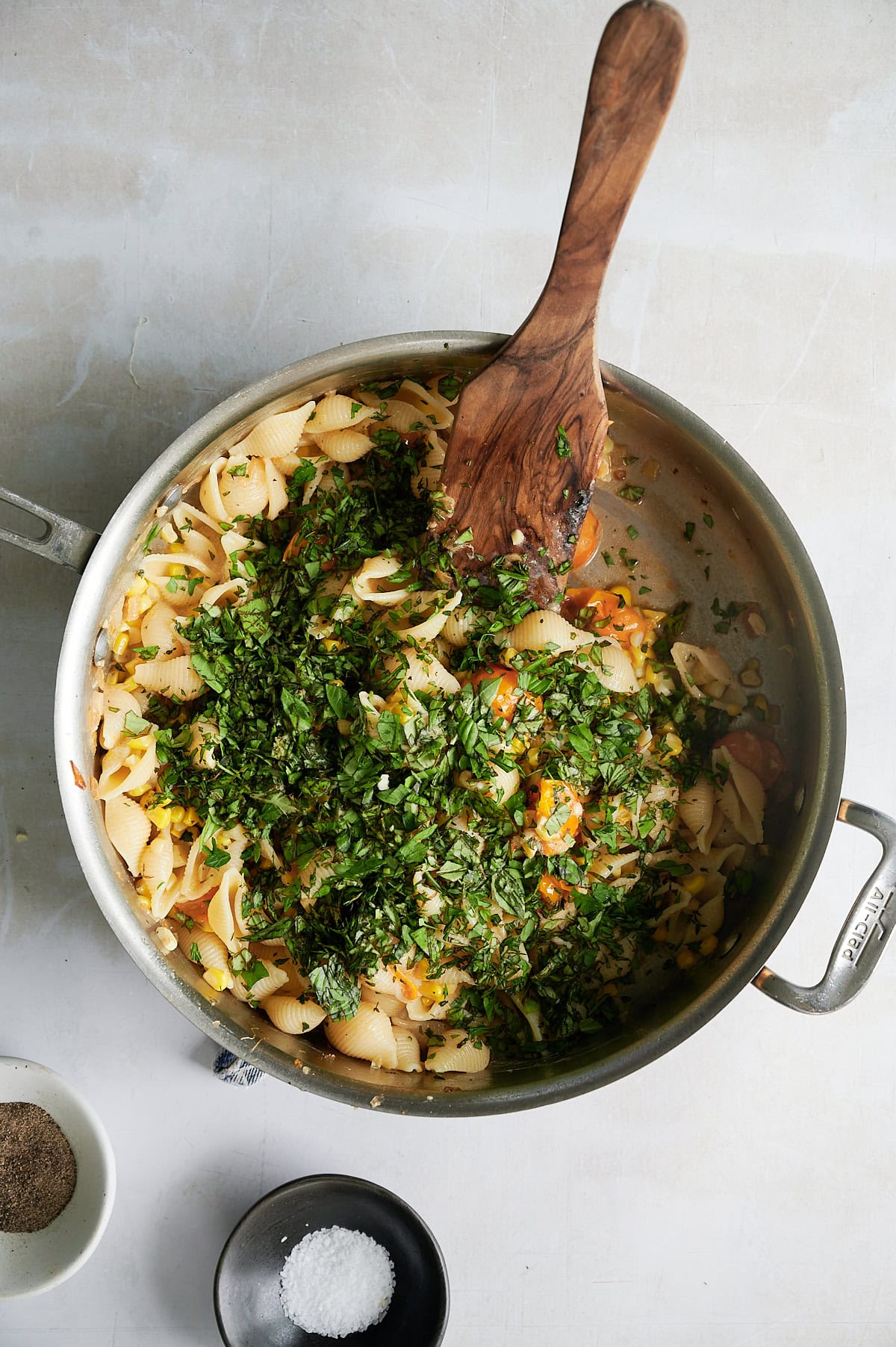 A pan of cherry tomato pasta with chopped herbs being stirred with a wooden spoon, next to small bowls of salt and pepper on a light surface.