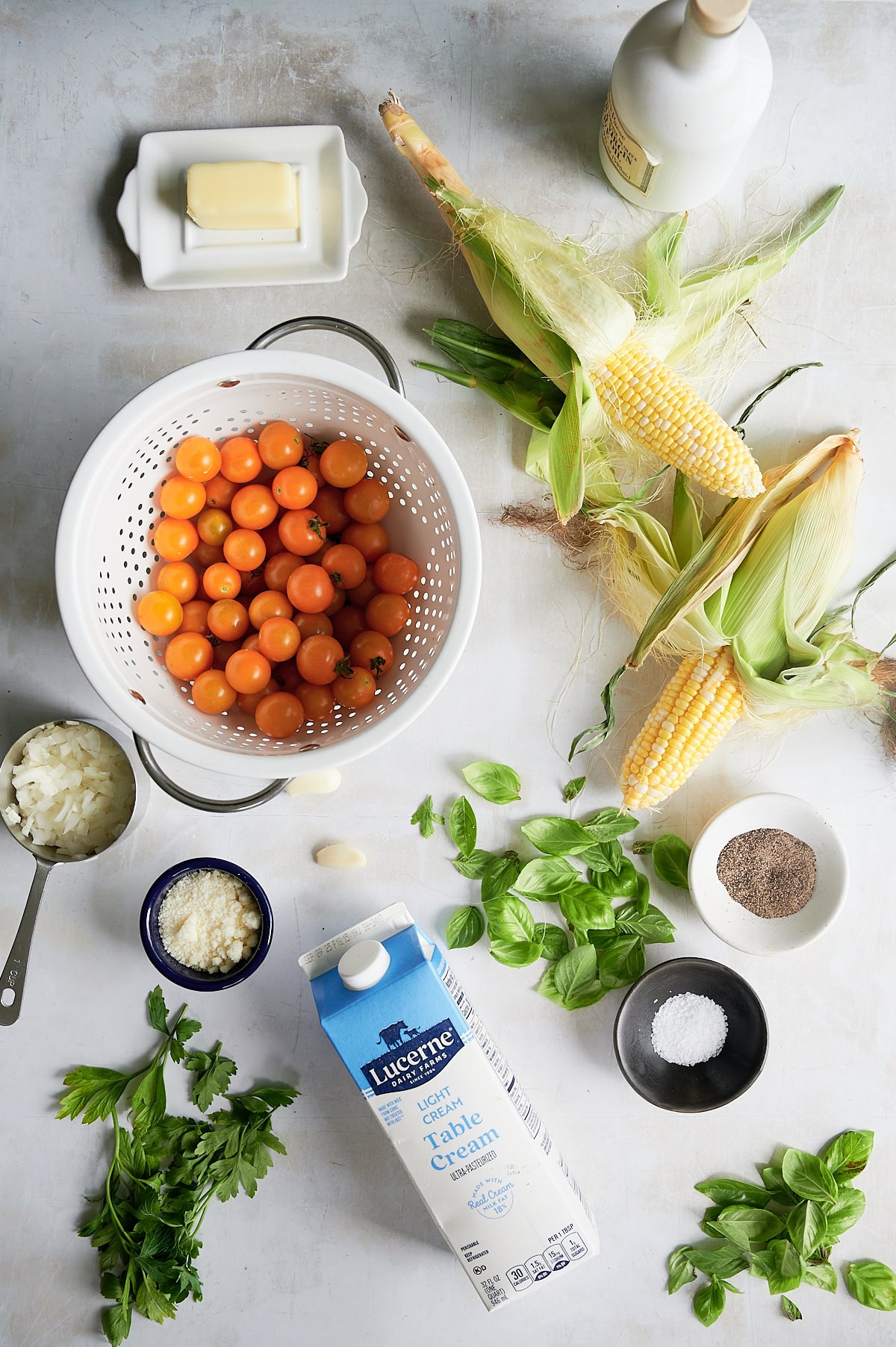 Overhead view of fresh ingredients for cherry tomato pasta, including cherry tomatoes, corn, basil, parsley, cream, butter, chopped onion, grated cheese, salt, pepper, and a bottle of oil on a light surface.