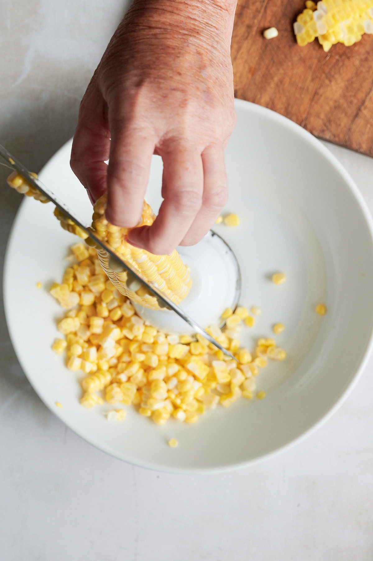 A hand cuts kernels off a corn cob into a white bowl, with diced corn pieces falling onto the plate.
