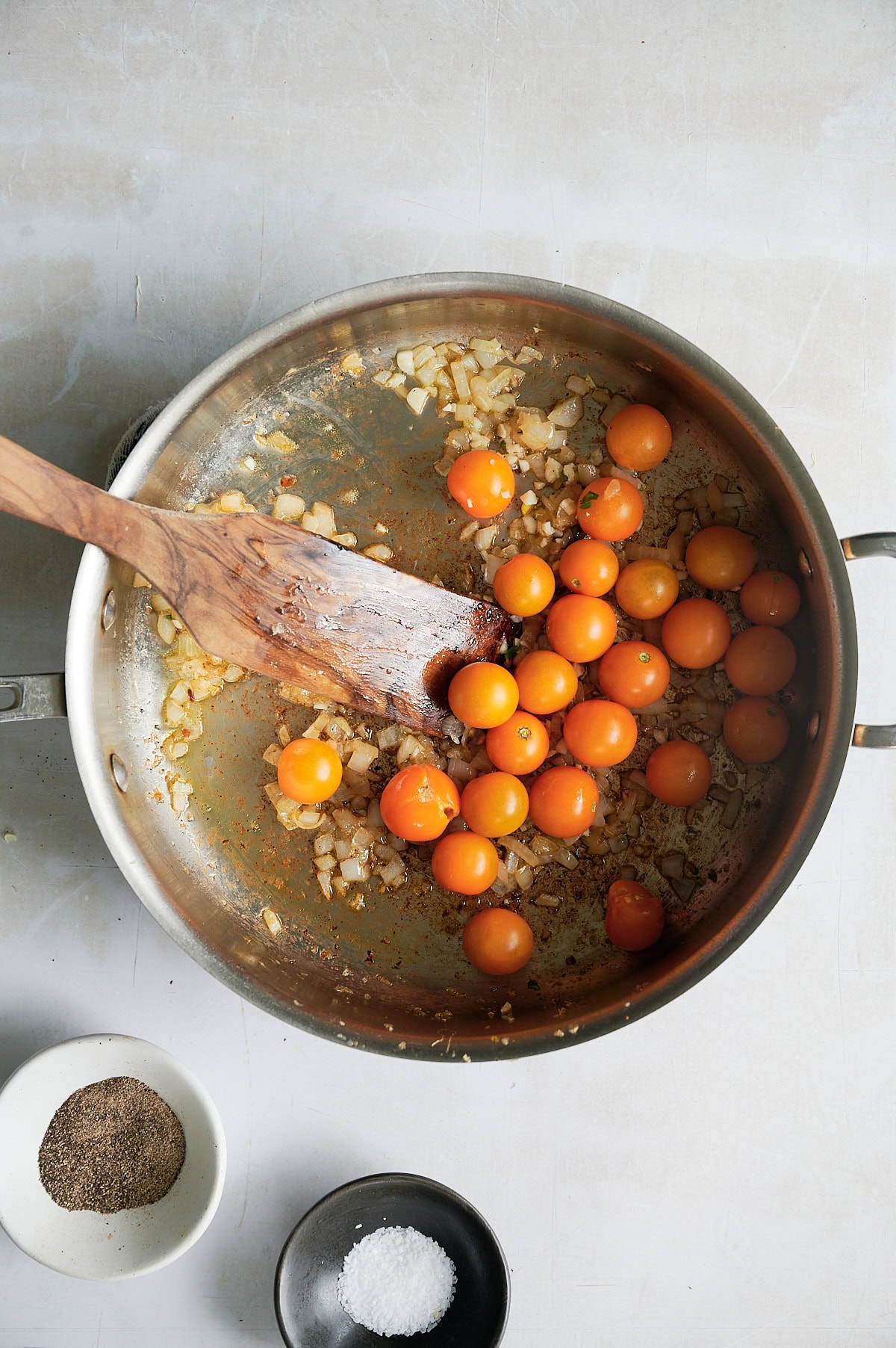 A stainless steel pan with sautéed onions and yellow cherry tomatoes, stirred with a wooden spatula; bowls of salt and pepper are nearby.